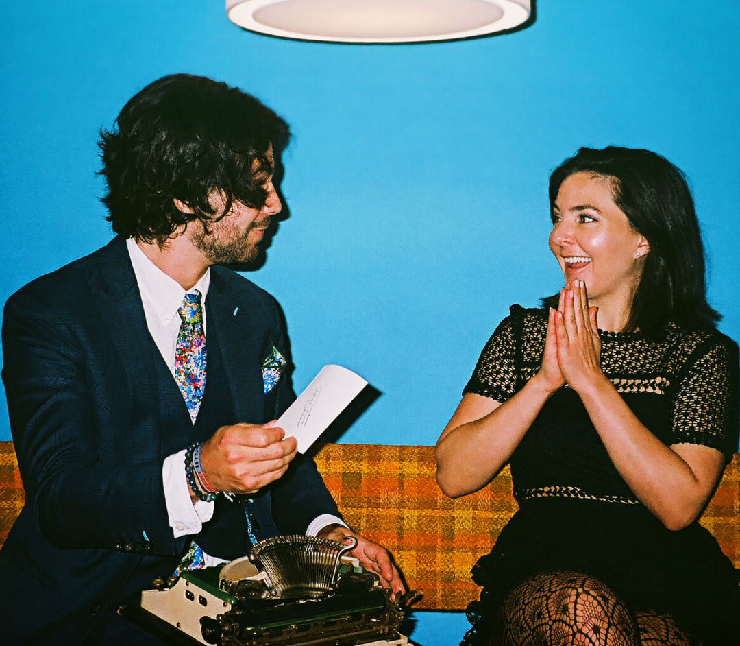 Man with typewriter and amused woman sitting against a blue background.