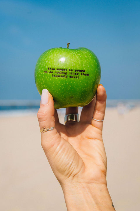 Green apple with haiku laser engraved on it held up with a coastline in the background.